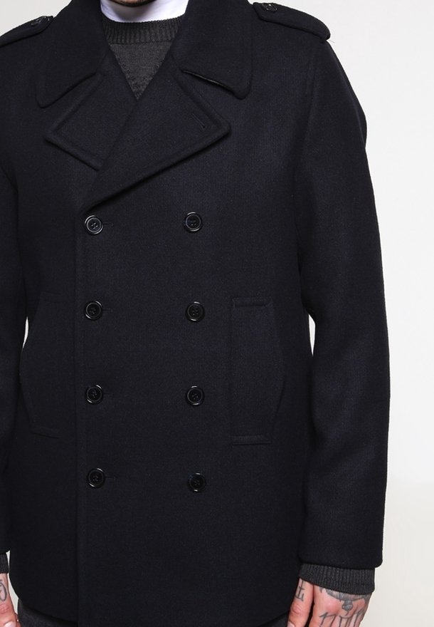 Mörkblå dubbelknäppt pilotjacka med sex svarta knappar, breda slag, axelkläppar och sidofickor på en person som bär en grå tröja.