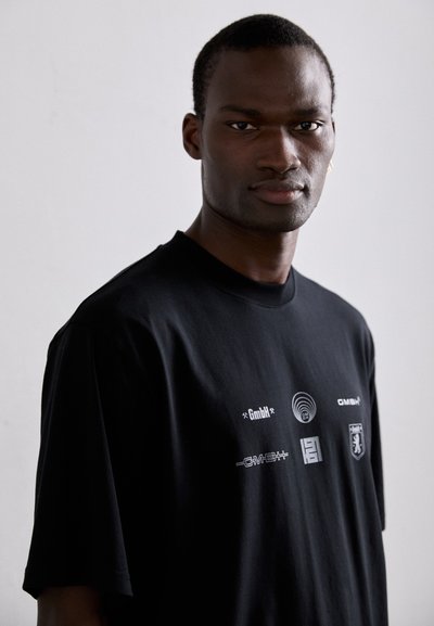 T-shirt en coton noir avec une coupe décontractée. Présente plusieurs impressions graphiques blanches sur la poitrine, y compris des logos et des symboles. Manches courtes.