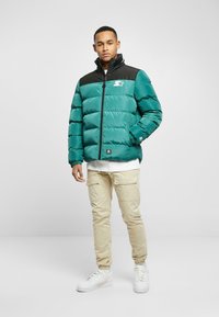 Teal puffer jacket with a black collar, zip closure, and logo detail. Worn over a white top and paired with beige cargo pants and white sneakers.