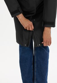 Manteau noir imperméable avec des poignets Velcro, devant ouvert, et fermetures à pression. Porté par-dessus un jean en denim bleu à large jambe avec une couture visible.