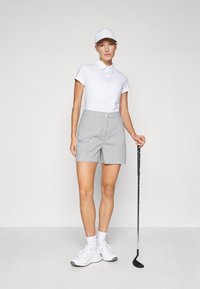 Camisa polo blanca con botones, pantalones cortos a cuadros en gris y blanco, y una gorra blanca. Sosteniendo un palo de golf, usando zapatillas blancas y calcetines.