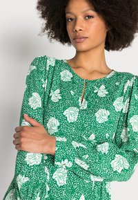 Vestido floral verde feito de tecido suave, com decote redondo, mangas bufantes e um detalhe em forma de chave na gola. Accentado com flores brancas.