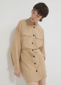 Beige Hemdblusenkleid mit langenÄrmeln, zwei Brusttaschen und schmaler Taille. Weicher Stoff und schwarze Knöpfe stehen im Kontrast zu der neutralen Farbe.