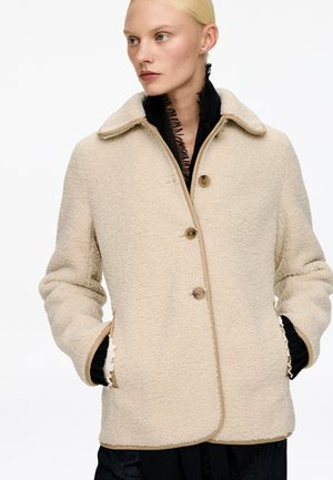 Beige Fleecejacke mit Kragen, Knopfleiste und Seitentaschen. Verfügt über kontrastierende braune Akzente und Textur für Wärme und Komfort.