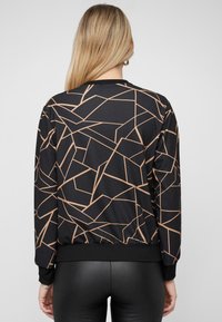 Blonde Frau, die sich abwendet und ein schwarzes Langarmoberteil mit beigen geometrischen Linien sowie schwarze enge Hosen trägt, vor einem schlichten Hintergrund.