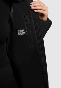 Sort pufferjakke med quiltet tekstur, sidelomme med lynlås og logo patch. Glat materiale, lige snit og tætsiddende pasform.