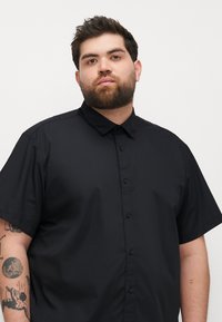 Zwarte shirt met korte mouwen en een button-down kraag, gemaakt van een gladde stof. Beschikt over een voorkant met knoopsluiting met zwarte knopen en heeft een relaxte pasvorm.