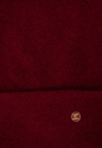 Rode gebreide beanie met een ribbelstructuur. Bevat een klein metalen logo label aan de zijkant, dat een cirkelvormig ontwerp toont.