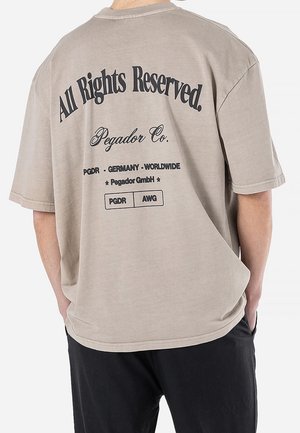 Ylimitoitettu taupenvärinen t-paita, jossa on lyhyet hihat ja musta teksti, mukaan lukien "All Rights Reserved" ja bränditiedot painettuna takana.
