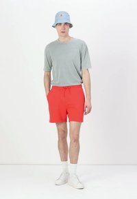 Maglietta grigia a maniche corte, pantaloni corti rossi con cordoncino, calzini bianchi alla caviglia e sneakers bianche. Cappello a secchiello blu in testa. Sfondo neutro.