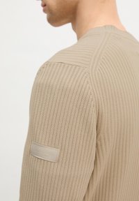 Pull en tricot beige texturé porté par une personne, montrant des coutures détaillées et une étiquette de marque en tissu rectangulaire sur le haut de la manche.