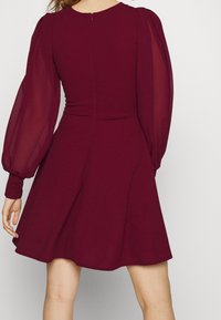 Kvinna i en vinröd klänning med genomskinliga puffärmar och flagad kjol, sedd bakifrån mot en neutral bakgrund.