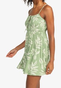 Vihreä mekko, jossa on kapeutettu vyötärö, valkoinen lehtikuvio, säädettävät olkaimet ja solmun etuosassa. Kangas vaikuttaa pehmeältä ja sillä on lumoava siluetti.