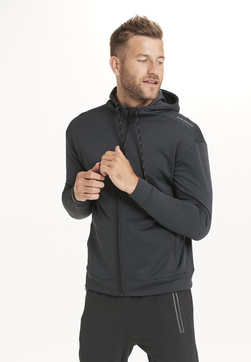 Musta vetoketjullinen huppari, jossa on teksturoitu kangas, säädettävät nyörit ja napakka leikkaus. Siinä on logo olkapäällä ja ribbireunat hihansuissa.