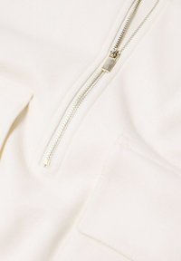 Prenda de vestir blanca con cremallera metálica, textura suave y un bolsillo con costuras. Diseño sencillo sin patrones ni detalles.