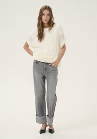 Suéter blanco de punto con un patrón curvado, combinado con jeans grises holgados que presentan puños deshilachados y zapatos de tacón negros. Fondo liso.