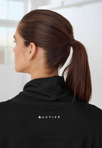 Haut de sport noir avec un col montant, arborant un petit logo blanc "ACTIVE" sur le haut du dos. Texture lisse et design ajusté.