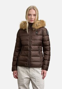 Giubbotto imbottito marrone con cappuccio foderato in pelliccia, chiusura con zip e design trapuntato. Presenta una toppa sulla manica e tasche laterali. Texture liscia.