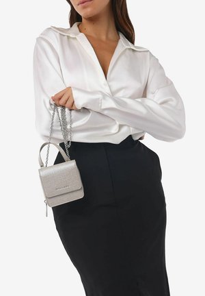 Silver glitter handbag with a rectangular shape, a chain strap, and a zip closure, held by a person wearing a white satin blouse and black skirt.