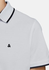 Polo shirt blanc en tissu texturé, doté d'un col rayé noir et d'un petit logo noir sur la poitrine. Les manches sont courtes.