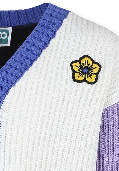 Pull en tricot blanc avec texture côtelée, accents violets et une broderie de fleur jaune bordée de noir sur l'épaule.
