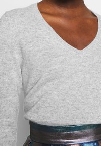 Gray v-neck sweater in soft knit with long sleeves, paired with a shiny teal and bronze waistband. Smooth texture, relaxed fit.