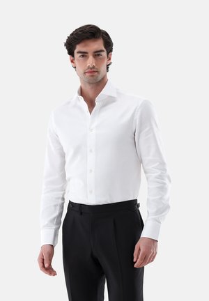 Uomo con capelli scuri che indossa una camicia bianca aderente infilata in pantaloni formali neri, in piedi contro uno sfondo chiaro e uniforme.