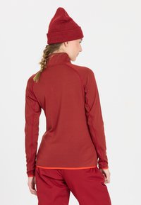 Rotes Langarmoberteil mit hohem Kragen, aus strukturiertem Stoff, mit kontrastierendem orangefarbenem Stich und tailliertem Design, kombiniert mit roten Hosen.