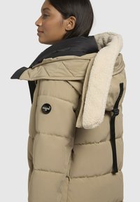 Bežs pūkains apģērbs ar kviltētu tekstūru un melniem akcentiem. Iezīmēts ar kažokādas odere cepuri un logo uzplāksteri uz piedurknes.