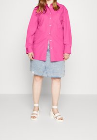 Vrouw in een felroze overhemd met knopen, een lichtblauwe denim rok met een versleten zoom en witte hoge hakken, staand op een effen achtergrond.