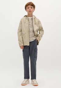 Giacca leggera beige con cappuccio, t-shirt a righe, pantaloni cargo blu scuro con tasche laterali e sneakers beige con accenti chiari.