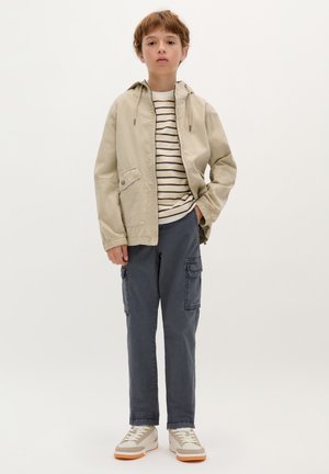 Veste légère beige avec capuche, t-shirt rayé, pantalon cargo bleu marine avec poches latérales, et baskets beiges avec des accents clairs.