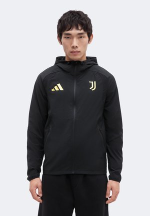 Junger Mann trägt eine schwarze Kapuzenjacke mit Reißverschluss und gelben Adidas- und Juventus-Logos, steht vor einem einfachen hellen Hintergrund.