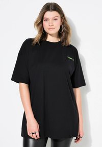 Černé oversized tričko vyrobené z bavlny, s malým zeleným logem na oblasti hrudníku. Krátké rukávy a kulatý výstřih.