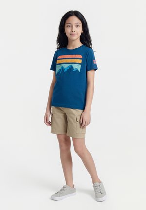 Dziecko z długimi ciemnymi włosami, ubrane w niebieski T-shirt z motywem górskim, beżowe spodenki cargo i szare trampki, stojące na białym tle.
