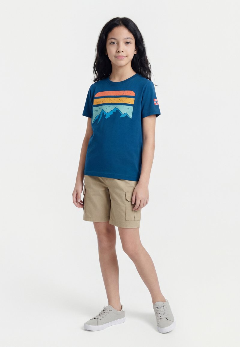 Παιδί με μακριά σκούρα μαλλιά, φοράει μπλε μπλουζάκι με σχέδιο βουνού, μπεζ cargo σορτς και γκρι αθλητικά παπούτσια, στέκεται μπροστά σε λευκό φόντο.