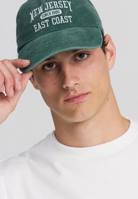 Cappello di cotone verde con la scritta "NEW JERSEY EAST COAST" ricamata in bianco, indossato da una persona con pelle chiara, abbinato a una camicia bianca.