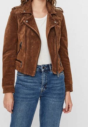 Frau trägt eine braune Wildleder-Bikerjacke über einem weißen Top und blauen Jeans, dargestellt von den Schultern bis zu den Hüften vor einem einfarbigen Hintergrund.