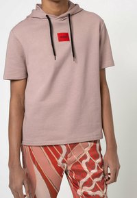 Persona che indossa un hoodie corti a maniche mauve con lacci neri e logo rosso, abbinato a pantaloni a fantasia rossi e beige.