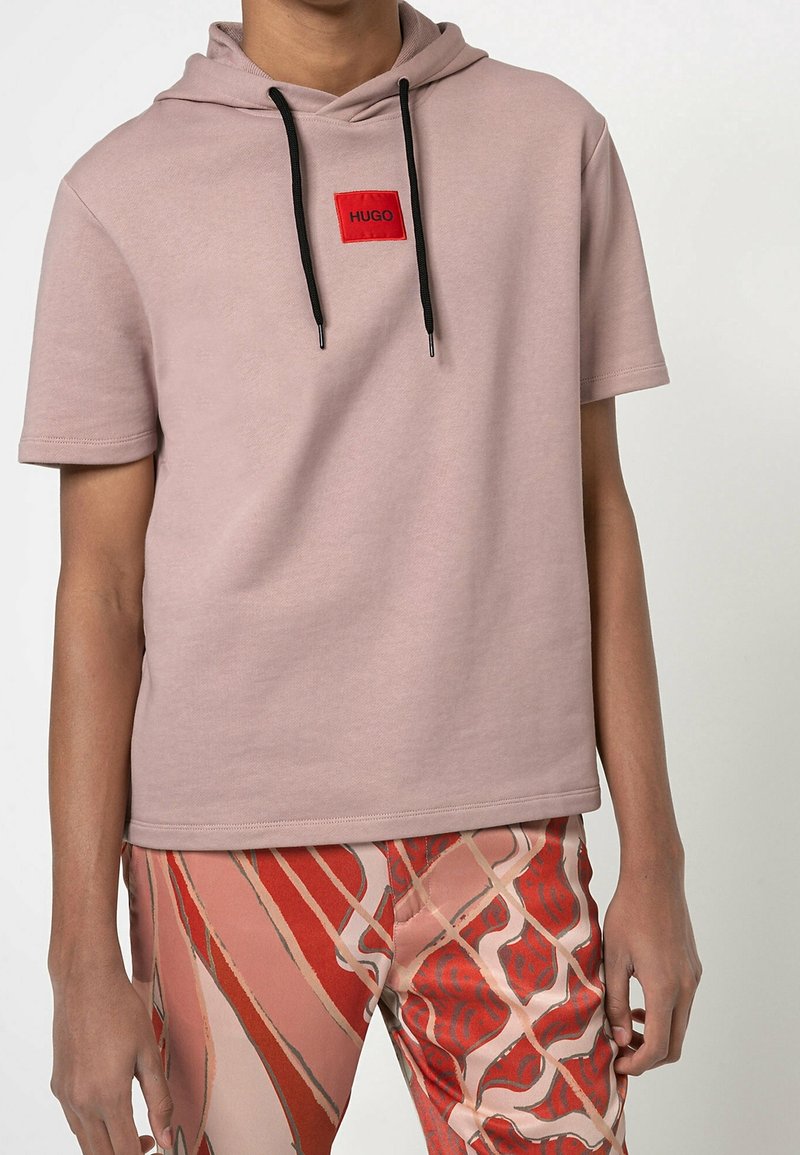 Persona che indossa un hoodie corti a maniche mauve con lacci neri e logo rosso, abbinato a pantaloni a fantasia rossi e beige.