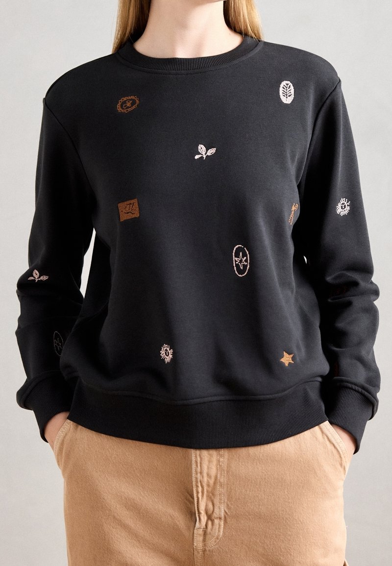 Personne portant un sweat-shirt noir avec de petits symboles brodés beige et marron clair, et un pantalon beige, les mains dans les poches, sur un fond uni.