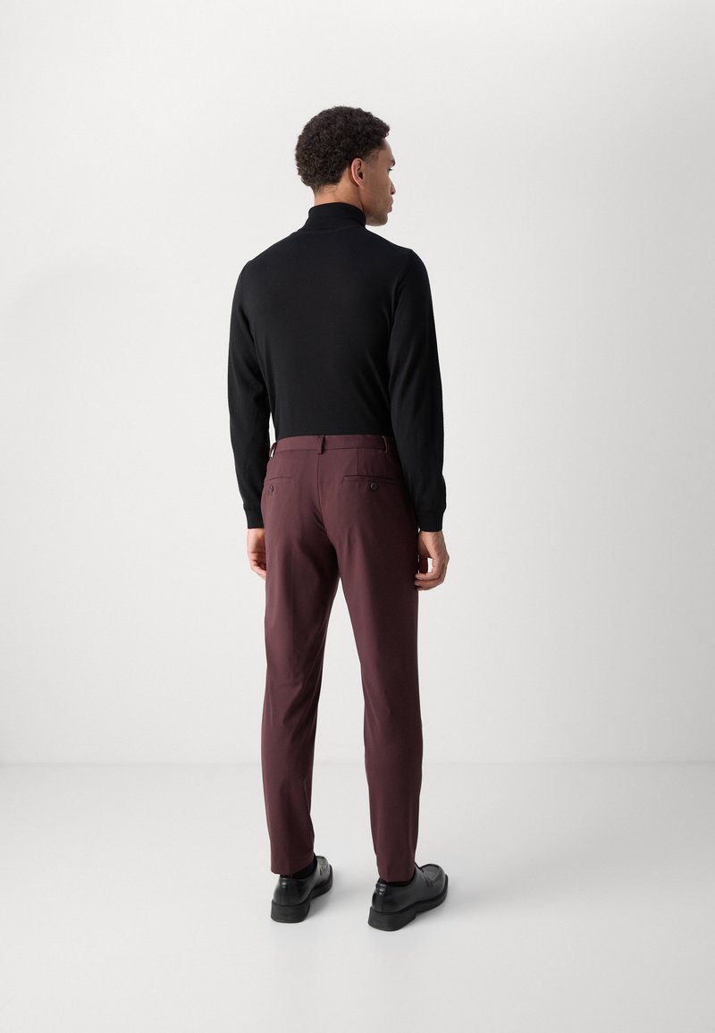 Schwarzes, hochgeschlossenes Langarmoberteil, kombiniert mit burgunderfarbenen Hosen. Die Hosen verfügen über Gesäßtaschen und einen maßgeschneiderten Schnitt. Schwarze Schuhe runden das Outfit ab.