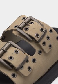 Μπεζ λουράκι σανδάλι με διπλή μεταλλική αγκράφα και πολλαπλές μεταλλικές τρύπες σε μαύρη σόλα.