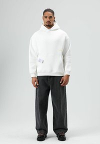 Sweat à capuche en coton blanc avec une poche avant et un logo jaune. Associé à un jean en denim noir ample et à des chaussures noires avec des lacets roses.