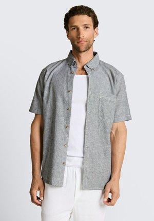 Homme aux cheveux courts et bouclés portant une chemise grise à manches courtes boutonnée sur un débardeur blanc et un pantalon blanc, debout devant un fond uni.