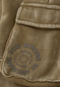 Khaki stof met een gestructureerde, geribbelde afwerking en een grote, reliëf circulaire graphic met de tekst "STAY STRONG BE AWESOME" op de zak.