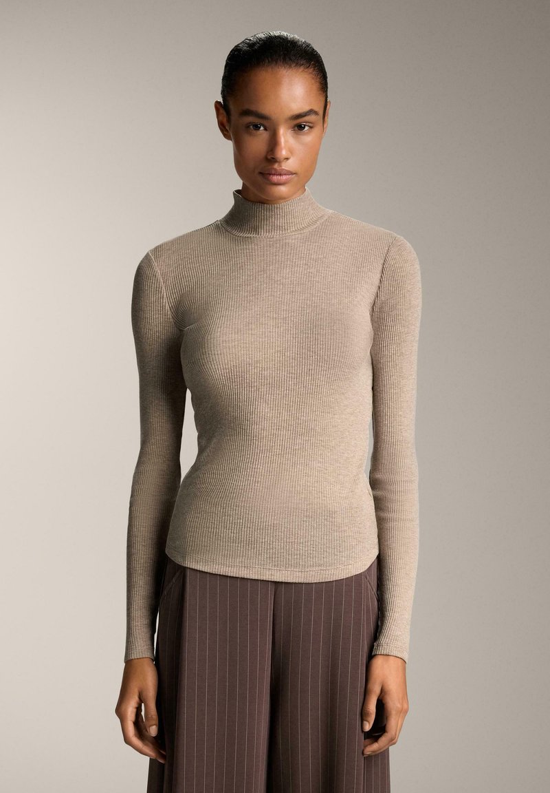 Kvinde med hår trukket tilbage, iført en beige ribbet turtleneck sweater og brune stribede bukser, der står foran en neutral baggrund.