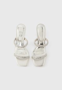 Ασημί σανδάλια ανοιχτής μύτης με δύο κυκλικούς ιμάντες διακοσμημένους με στρας και τακούνι block, με λείο μεταλλικό φινίρισμα.