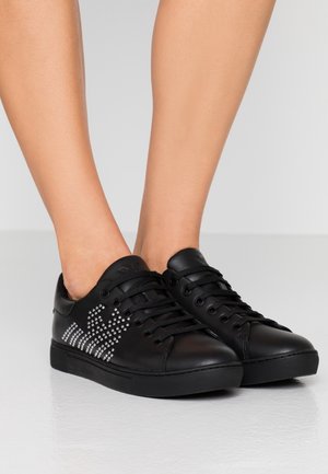 Sorte lavtop-sneakers med sorte snørebånd og sølvnagledesign på siden, båret på fødder mod en ensfarvet lys baggrund.