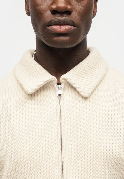 Κοντινό πλάνο ενός ατόμου που φοράει πλεκτό πουλόβερ σε κρεμ χρώμα με διπλωμένο γιακά και μπροστινό φερμουάρ μισάνοιχτο.